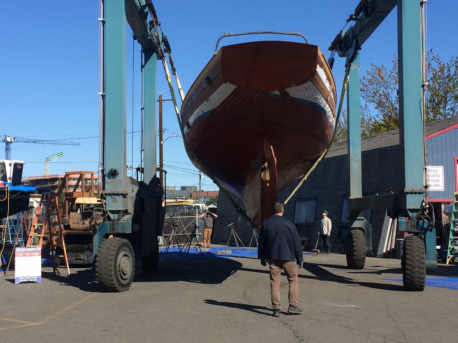 The classic 1946 Philip Rhodes-designed Merry Maiden is carefully hoisted by massive travel lift cranes at a Pacific Northwest boatyard, her wooden hull suspended between turquoise steel beams against a clear blue sky.
