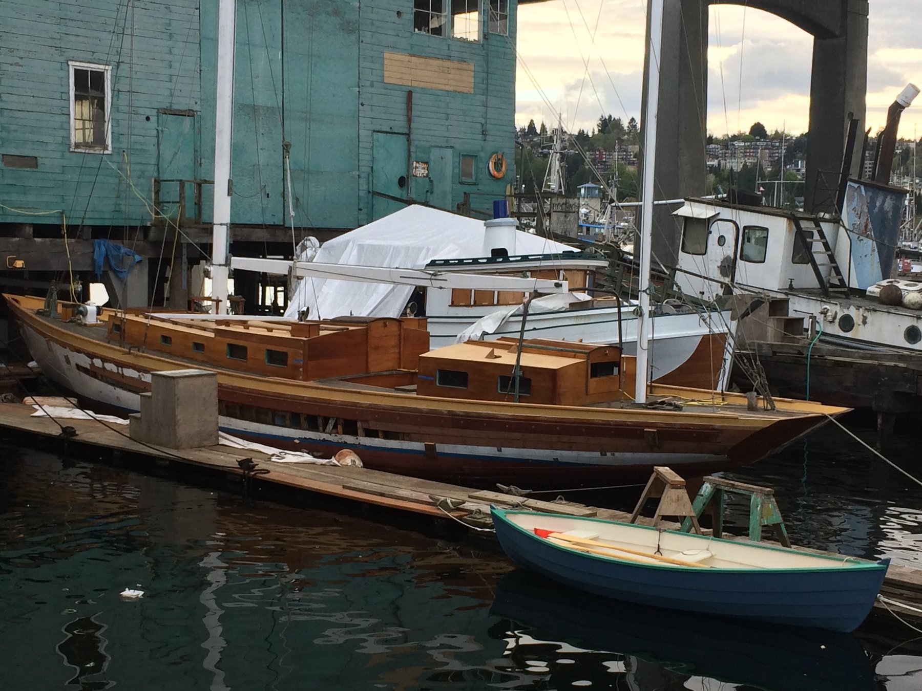 The classic wooden hull of Merry Maiden rests peacefully at dock, her traditional lines and warm timber construction a testament to Philip Rhodes' 1946 design, surrounded by the working vessels and industrial waterfront of what appears to be a Pacific Northwest marina.