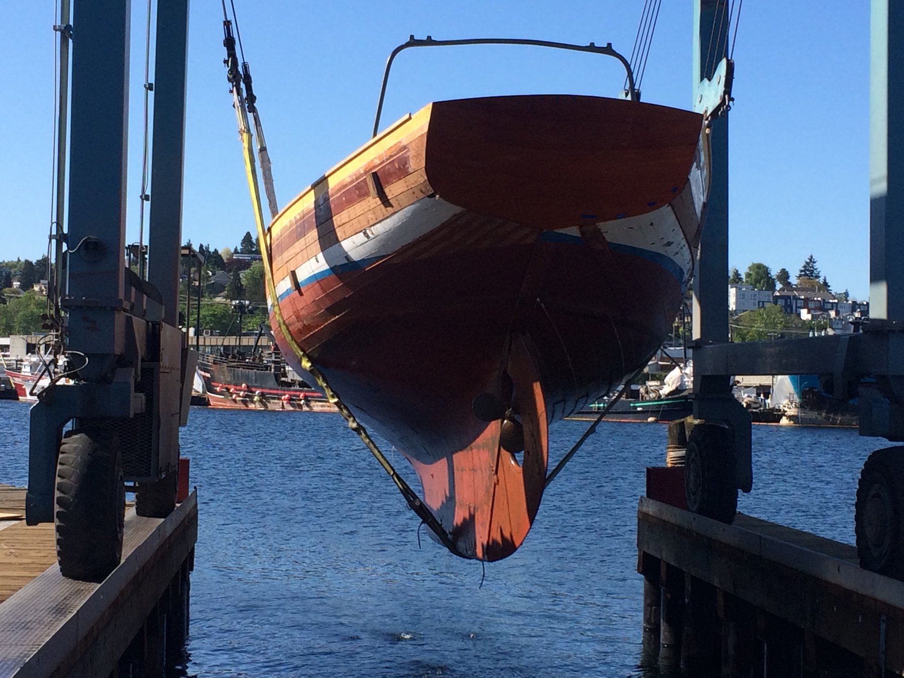 The Merry Maiden, a 1946 Philip Rhodes classic, hangs suspended between the boat lift's cradles in a gleaming moment of maintenance, her beautiful wooden hull and distinctive paint scheme fully exposed in the bright Pacific sunshine.