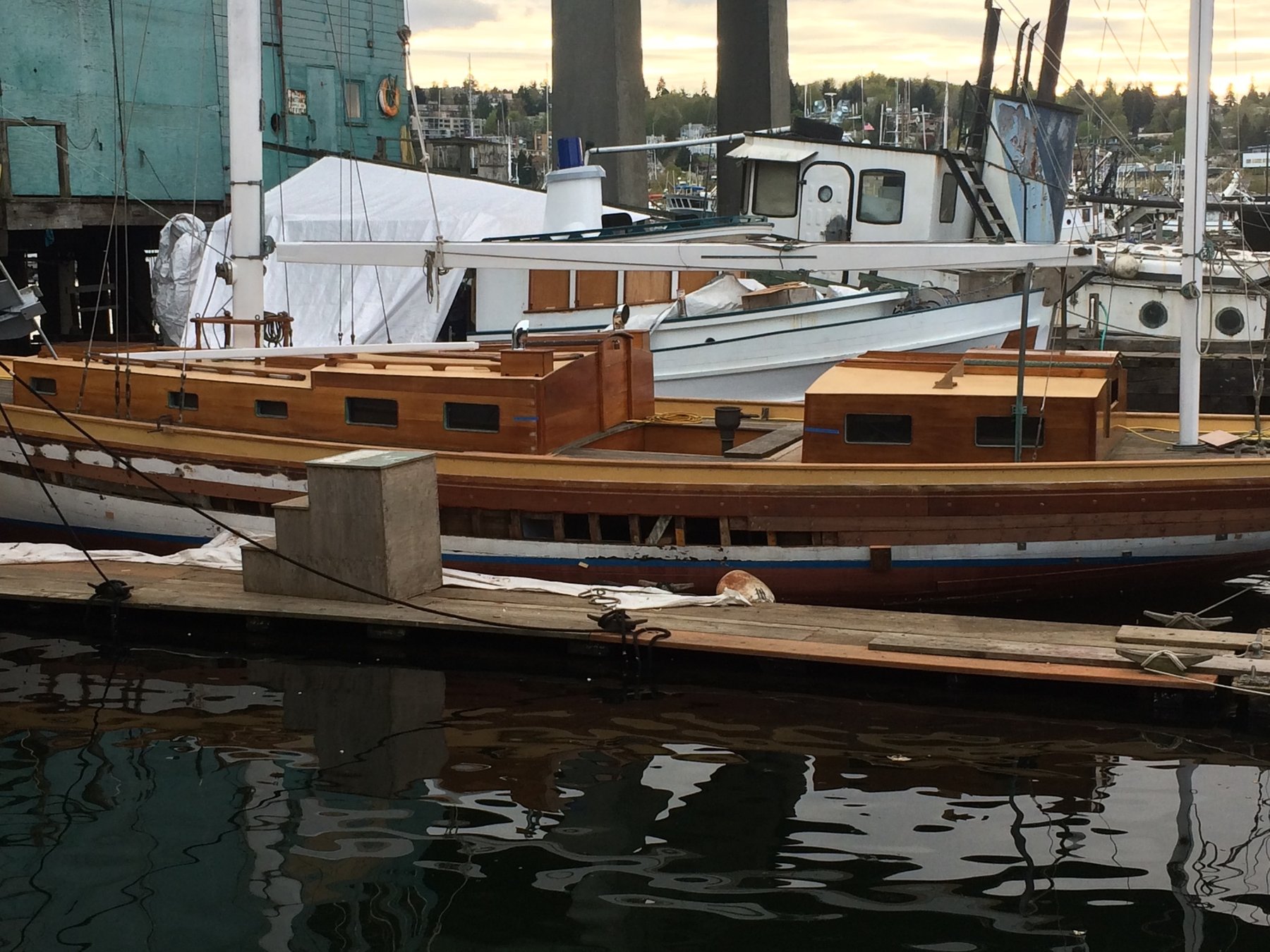 The classic wooden hull of Merry Maiden rests peacefully at the dock, her warm teak and mahogany tones reflected in the still water of the marina, with other vessels and forested shores visible beyond the pilings.