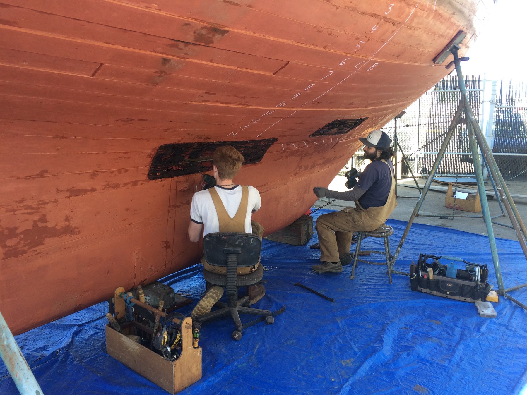 Two craftsmen work beneath the hull of the classic 1946 Philip Rhodes-designed Merry Maiden, carefully inspecting and documenting the wooden planking during restoration in the Puget Sound boatyard.