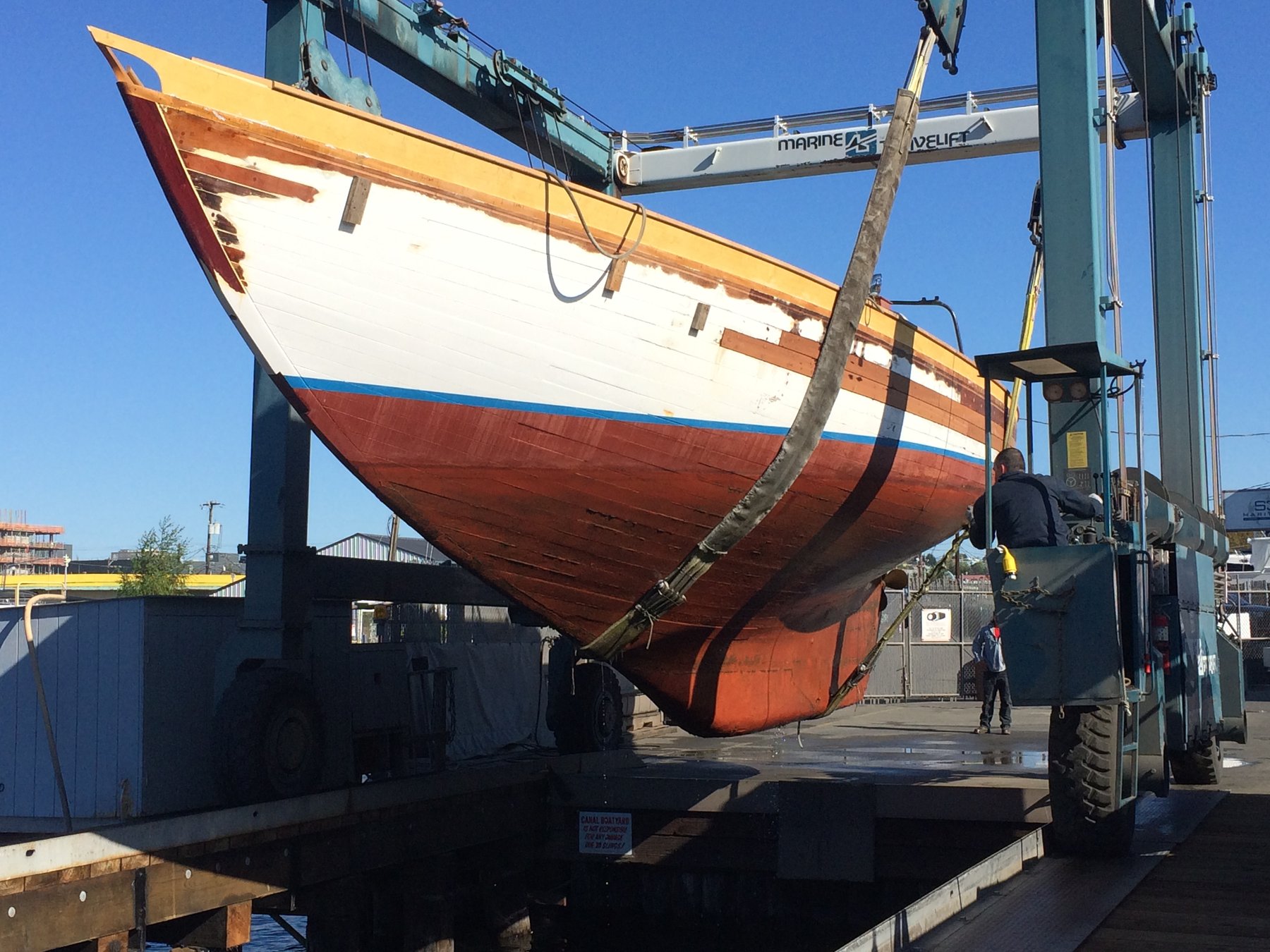 The Merry Maiden, a 1946 Philip Rhodes classic, hangs suspended in the travel lift at a Pacific Northwest boatyard, her iconic wooden hull and contrasting paint scheme on full display during haul-out.