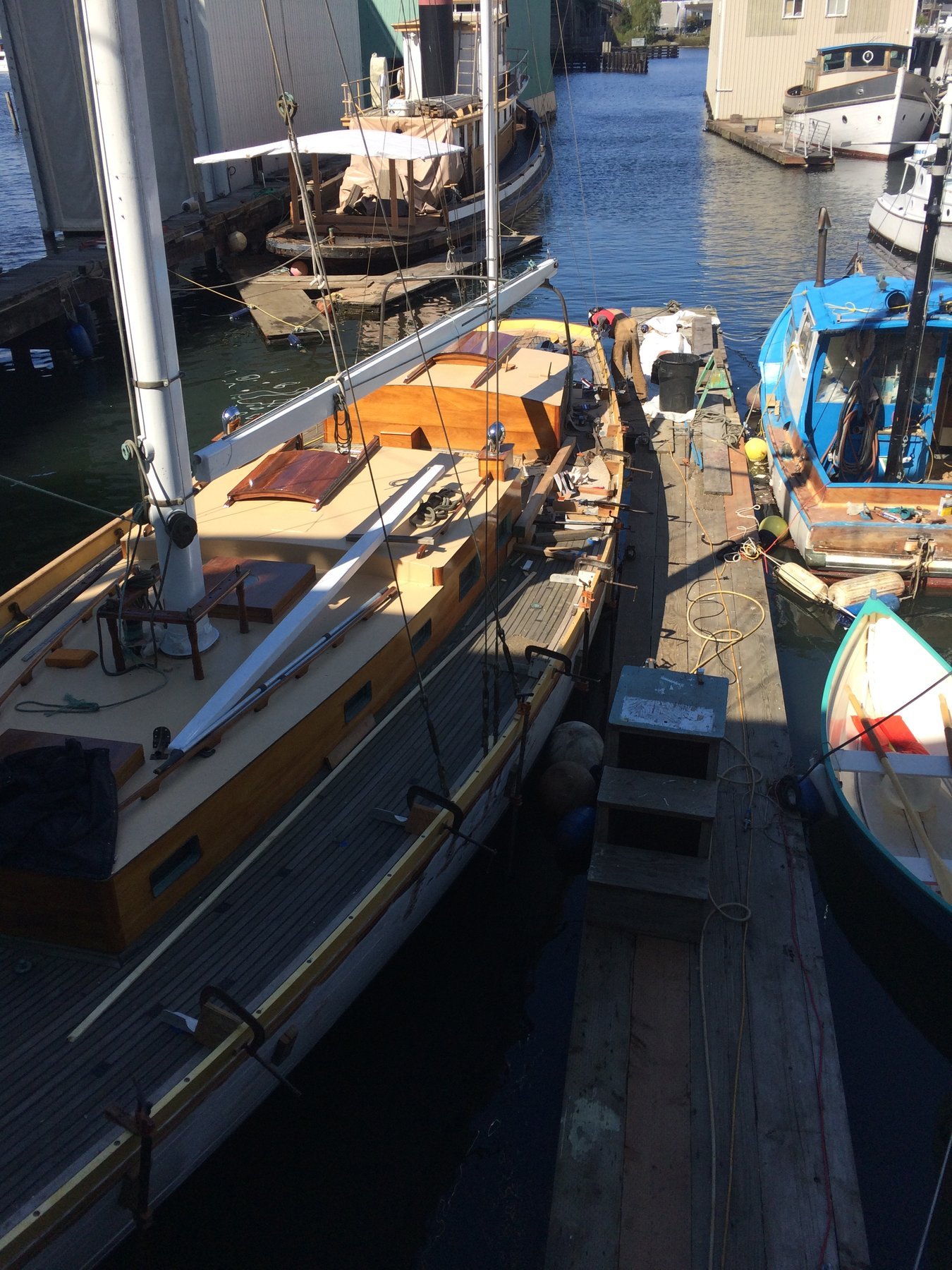 The classic 1946 Philip Rhodes-designed Merry Maiden rests at her mooring in Puget Sound, her cream and dark hull gleaming in spring sunlight alongside a working boatyard.