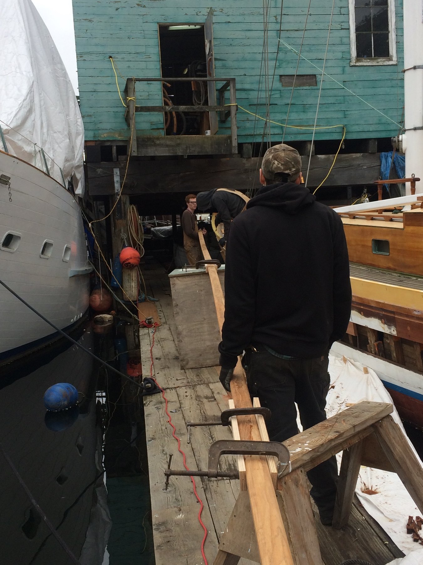 Craftsmen work on the wooden hull of the classic 1946 Philip Rhodes-designed Merry Maiden during restoration, with her sturdy frame and neighboring vessels visible in the boatyard.