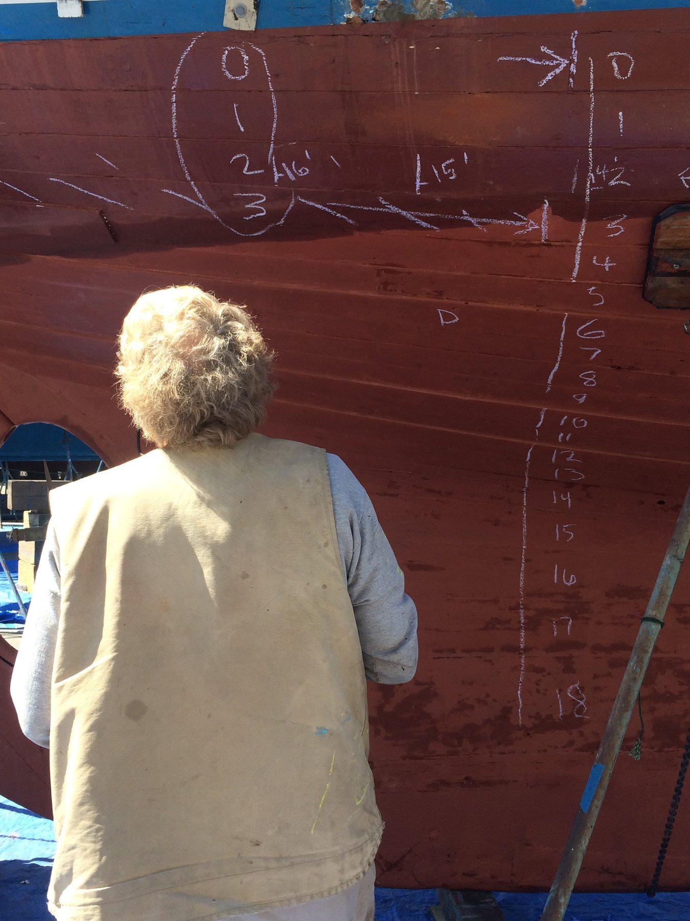 A crew member tends to canvas work on the classic 1946 Philip Rhodes-designed Merry Maiden, her weathered hull marked with measurements and notes during restoration or maintenance in Puget Sound.