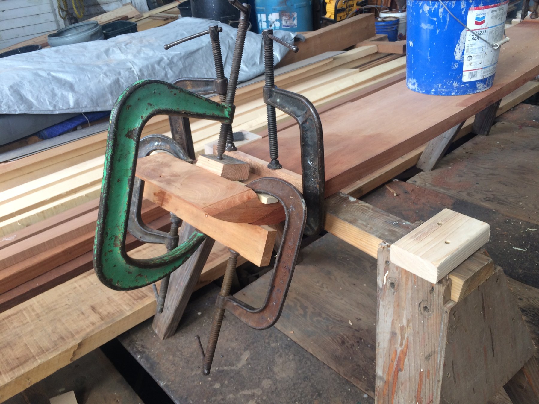 A vintage wooden clamp press grips a freshly milled timber in the boatyard, surrounded by stacked planking destined for the restoration of this classic 1946 yacht.