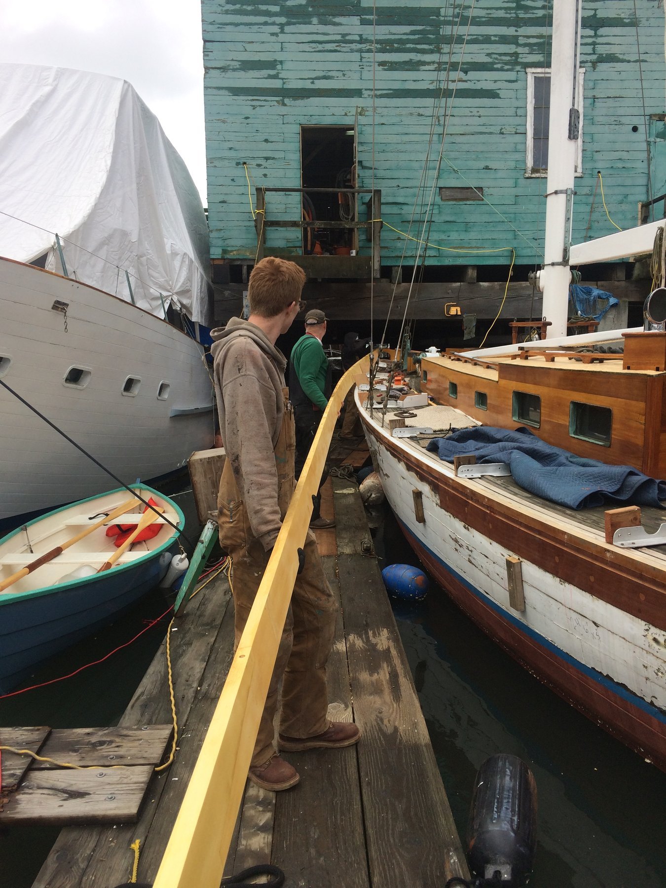 A crew member tends to the yellow spinnaker pole of the classic 1946 Philip Rhodes-designed Merry Maiden, moored alongside weathered pilings in Puget Sound with her graceful wooden hull and traditional rigging on full display.