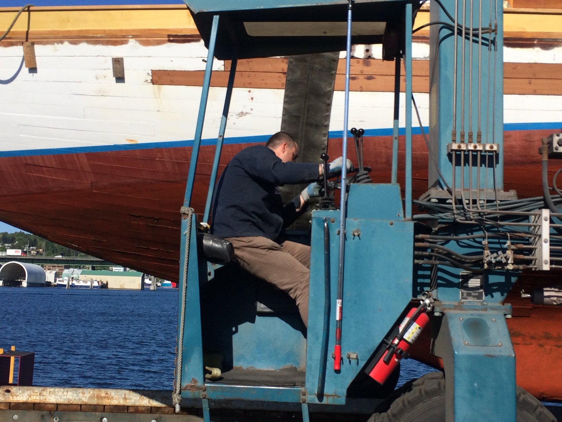 A crew member works on the hydraulic systems of the classic 1946 Merry Maiden while she rests in the boatyard, her distinctive red and white hull prominent against the water.