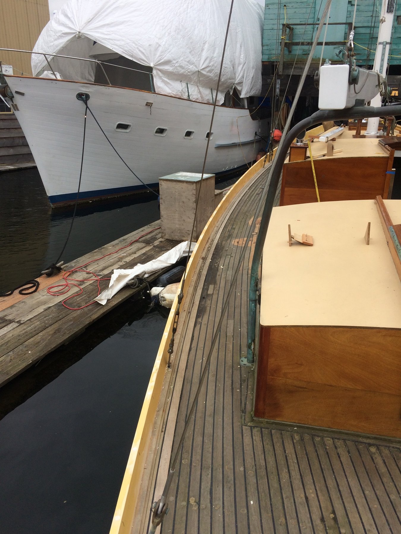 The classic 1946 Philip Rhodes design Merry Maiden rests at the dock, her weathered teak decks and wooden cabin sides showing the patina of decades, while a neighboring vessel's protective white cover stands sentinel nearby.