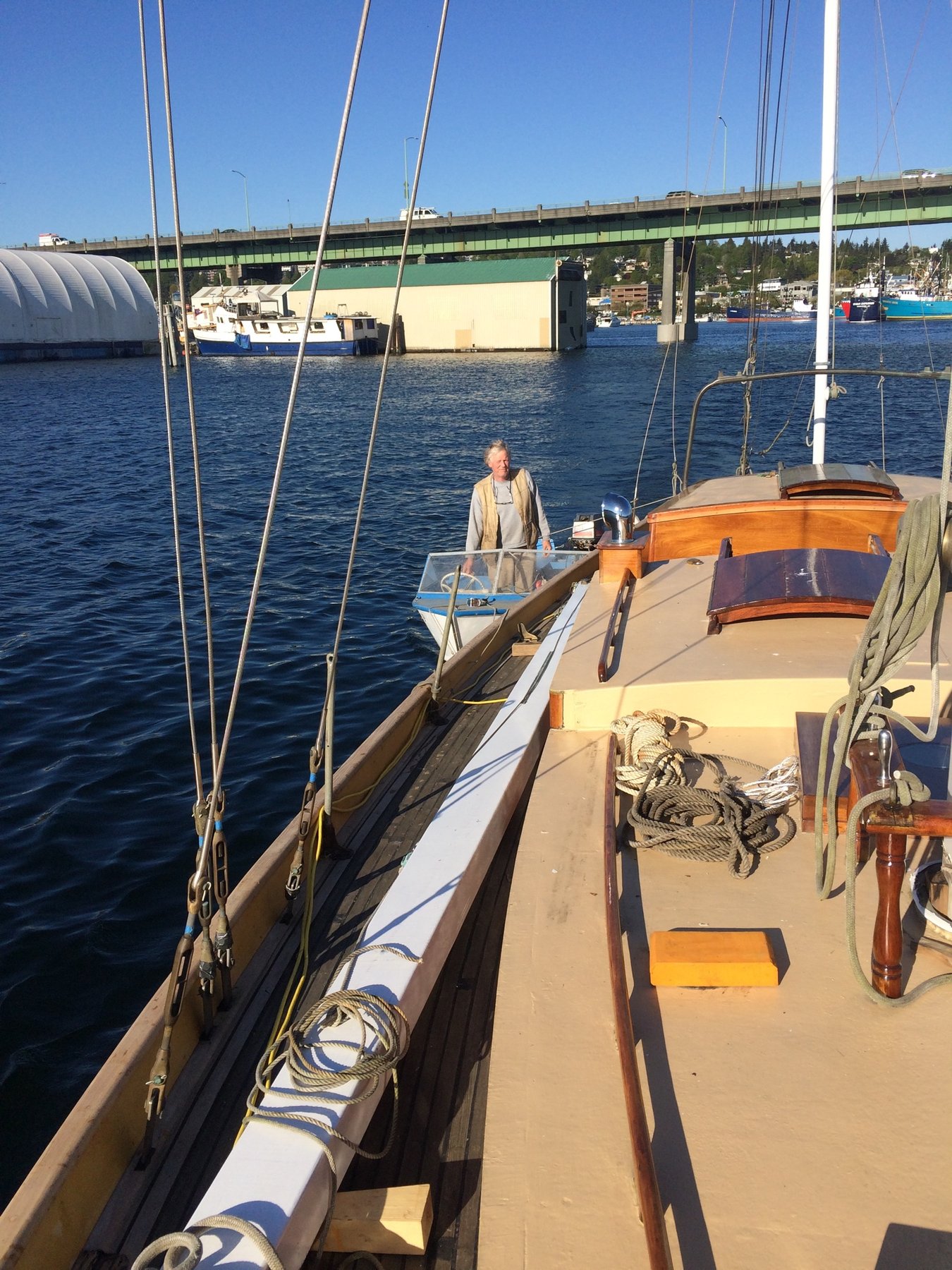 The classic 1946 Philip Rhodes-designed Merry Maiden rests at her moorings in a Pacific Northwest harbor, her teak deck and traditional wooden details gleaming under clear skies as a crew member tends to her rigging.
