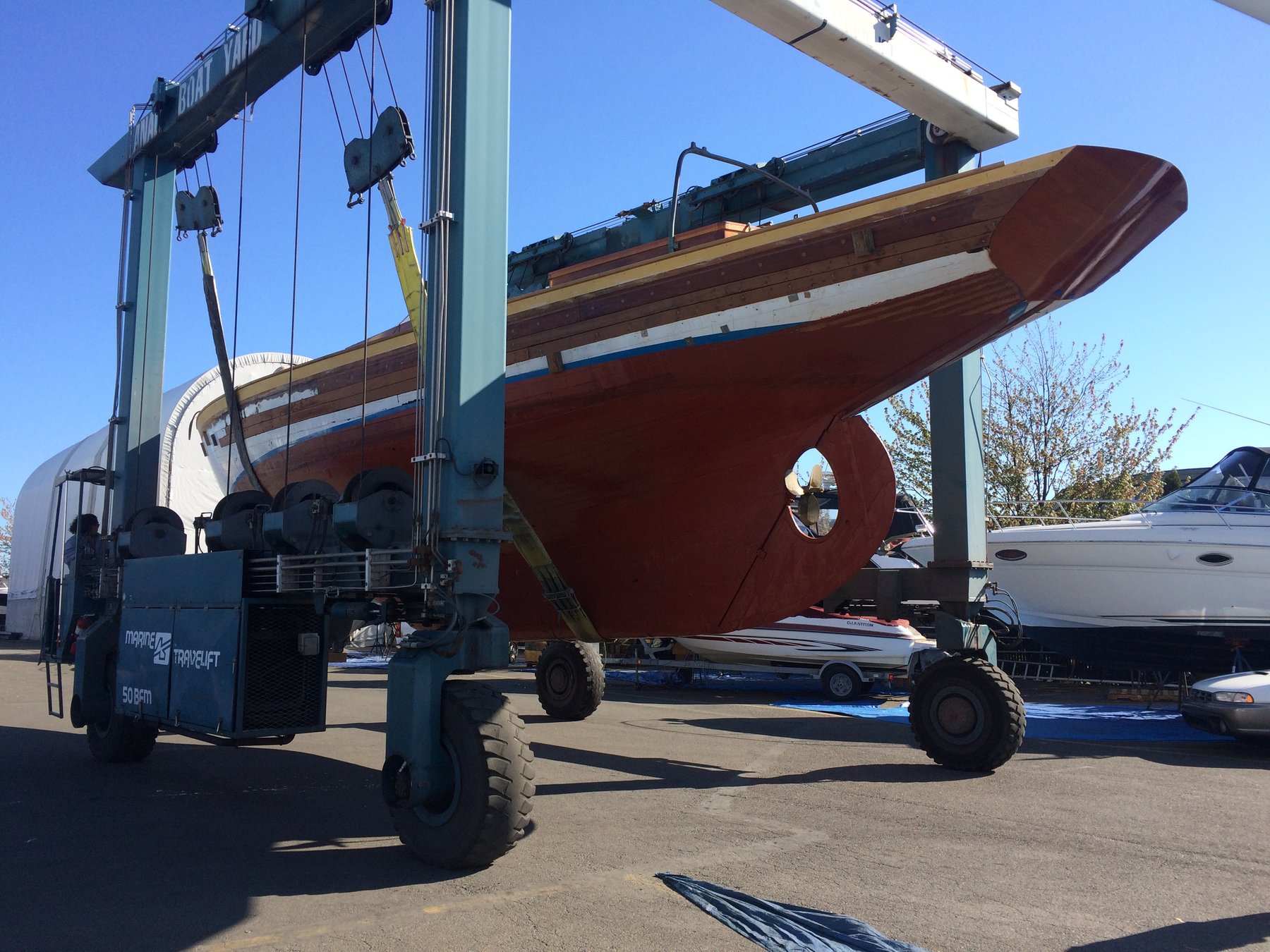 The classic 1946 Philip Rhodes-designed Merry Maiden is cradled by a massive travel lift at the Puget Sound boatyard, her elegant wooden hull and distinctive red boot stripe suspended against the clear Pacific Northwest sky.