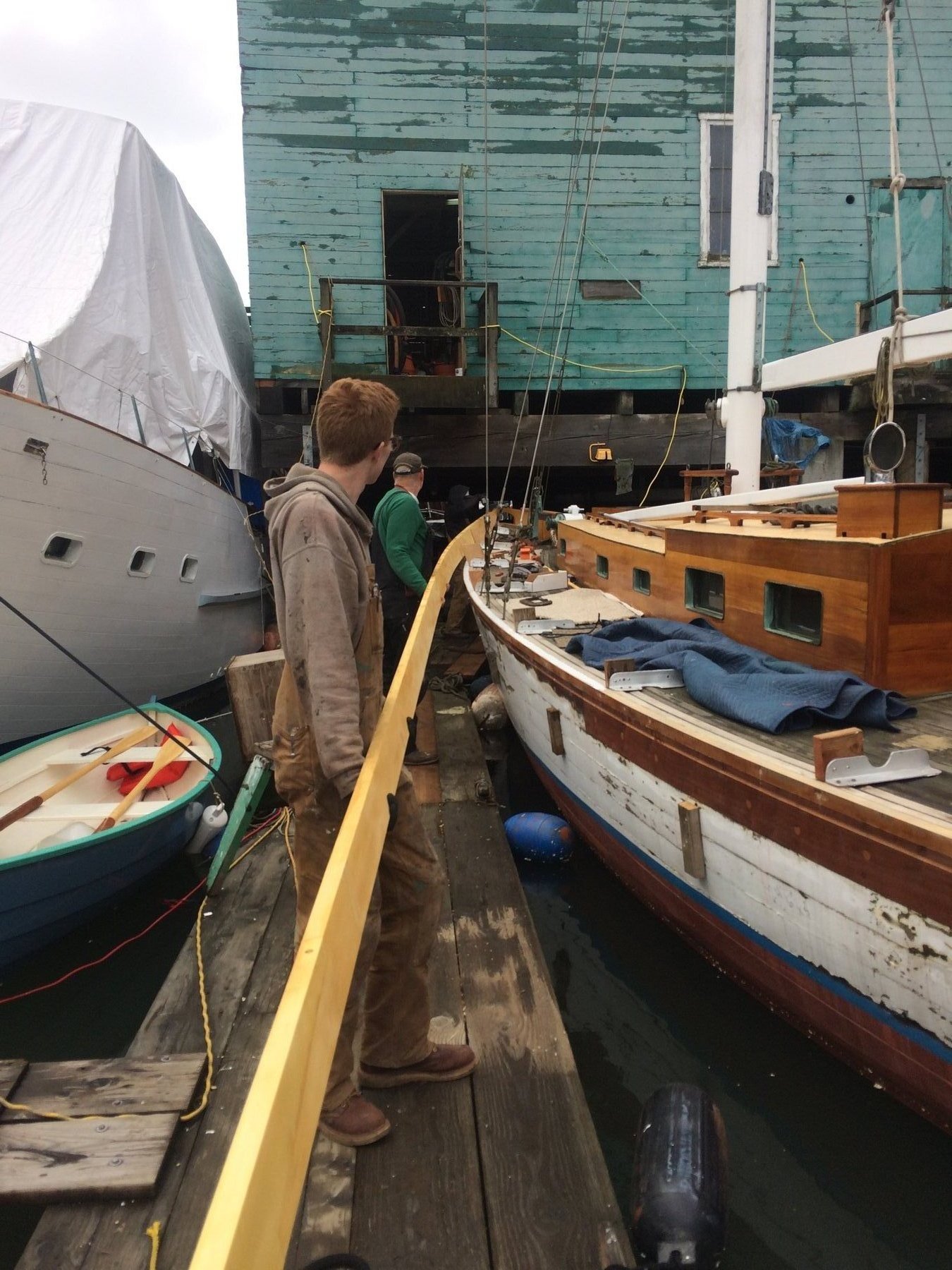 A crew member tends to the Merry Maiden's rigging at the dock, with the classic 1946 Philip Rhodes design showing her graceful lines and well-maintained wooden hull against the weathered boatyard backdrop of Puget Sound.