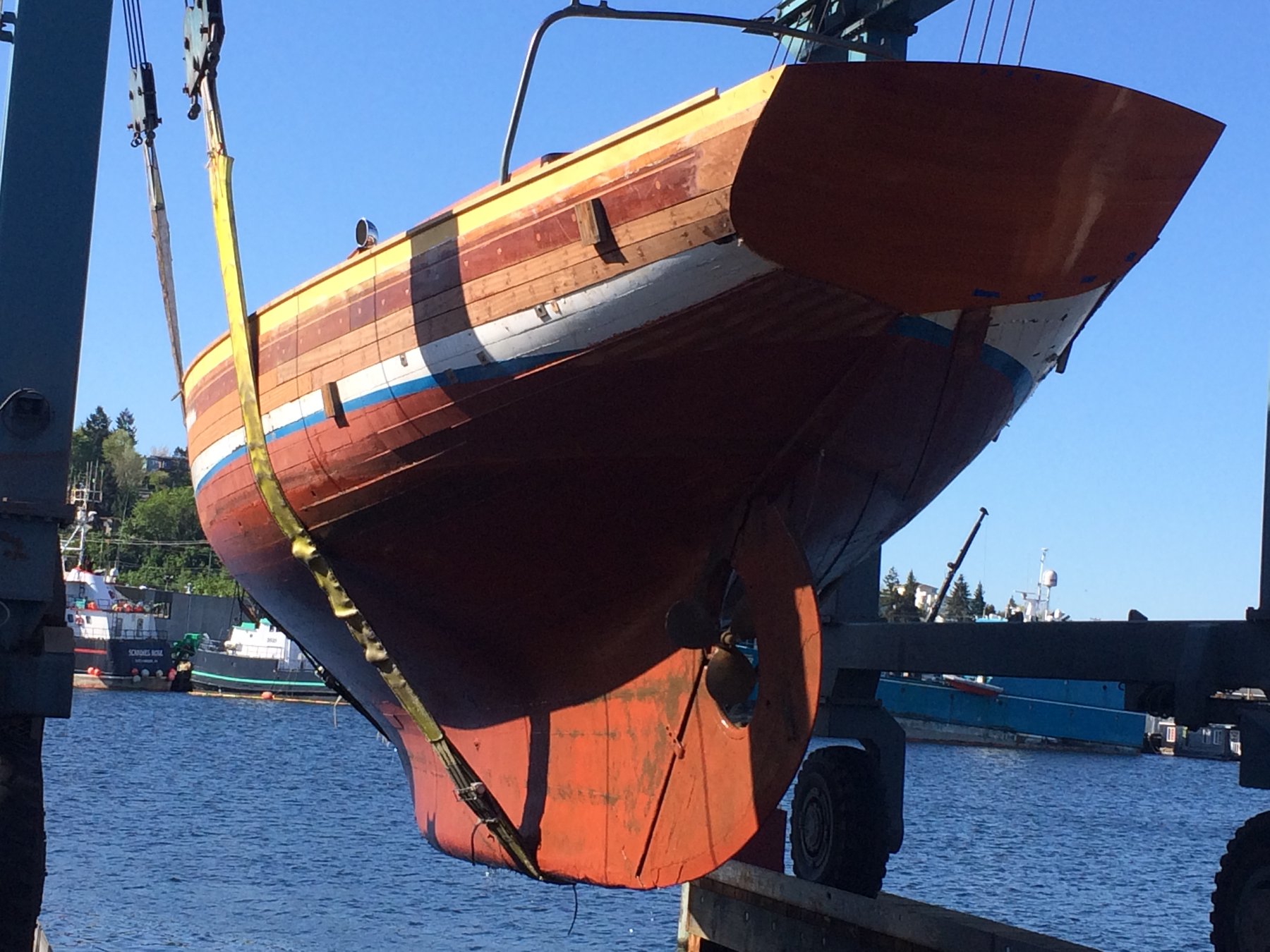 The Merry Maiden, a 1946 Philip Rhodes classic, hangs suspended from the travel lift in Puget Sound, her wooden hull and distinctive striped topsides gleaming in the spring sunshine during haul-out.