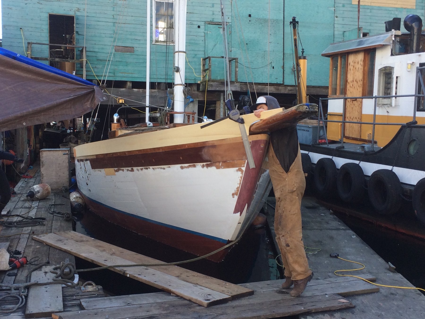 A worker tends to the classic lines of Merry Maiden during restoration work in the boatyard, her white hull and mahogany brightwork revealing both age and character from her 1946 launch.