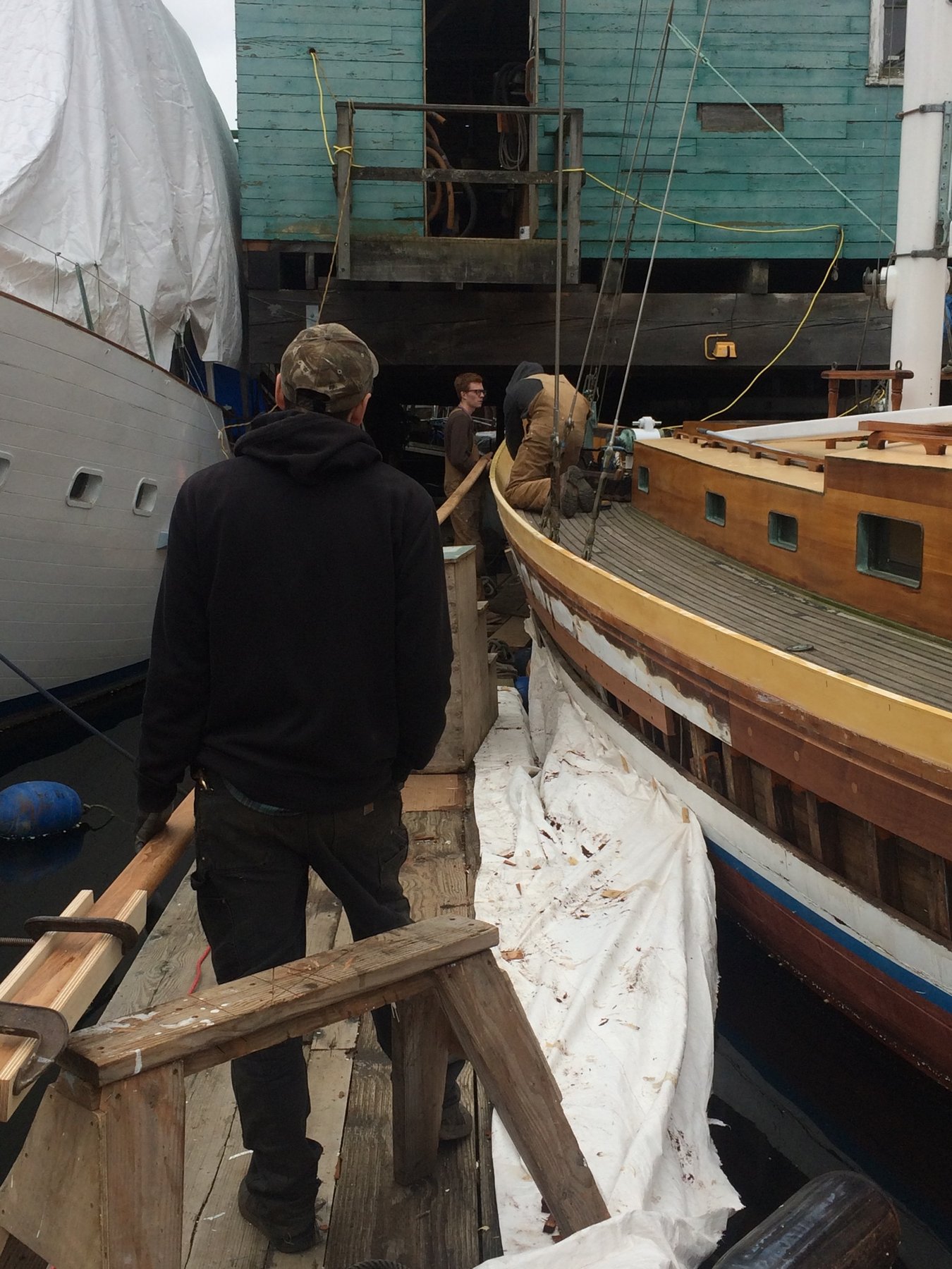 Craftsmen work on the wooden hull of the classic 1946 Merry Maiden during restoration, with protective coverings and scaffolding surrounding the historic 35-foot yacht in the boatyard.