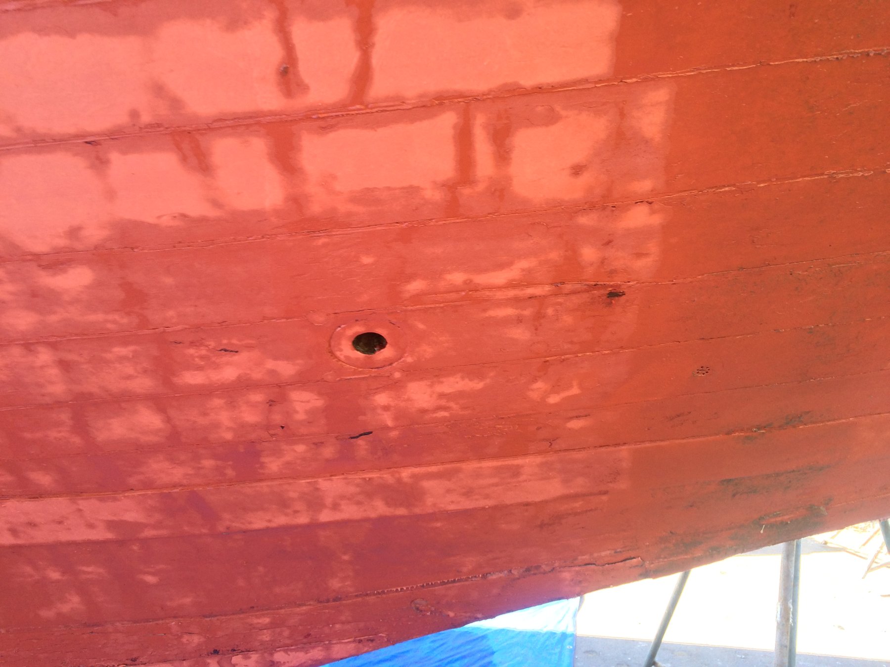 Overhead view of Merry Maiden's red-painted deck showing the patina of age and character of this 1946 classic, with shadows cast across the weathered wooden planking and a single through-hull fitting visible.
