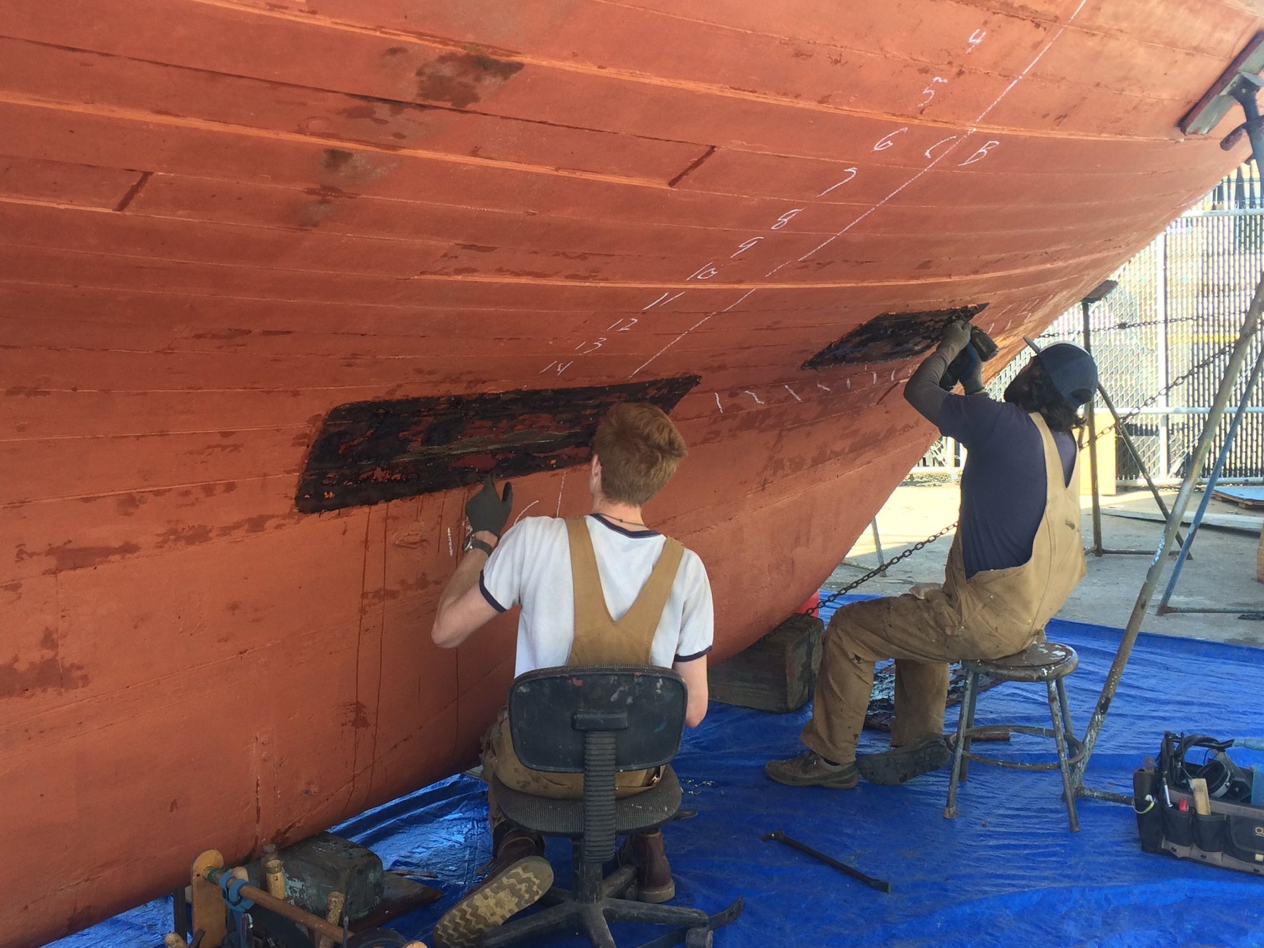 Two craftsmen work on the hull of the classic 1946 Merry Maiden, carefully scraping away old paint to reveal the wooden planking beneath in preparation for restoration.