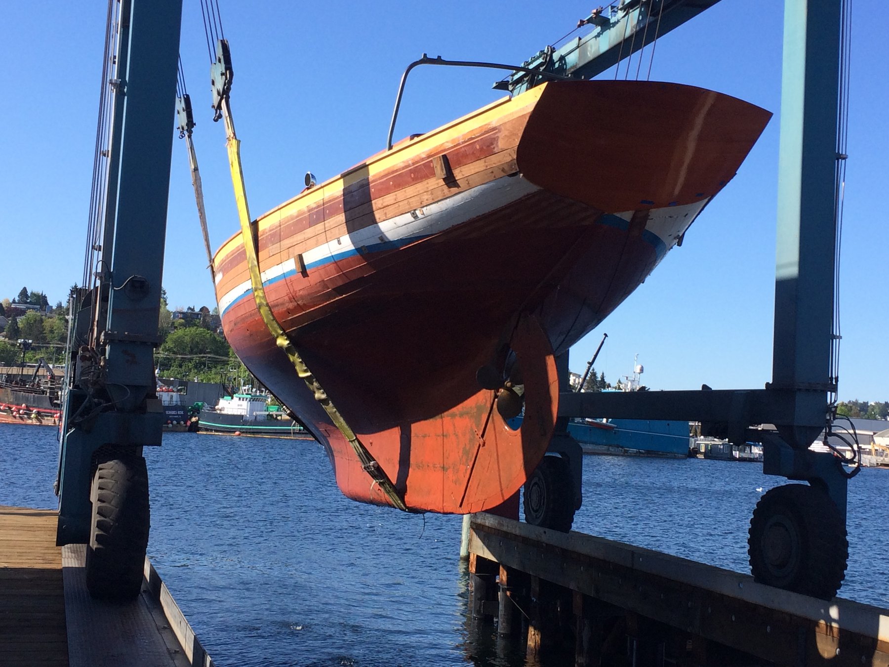 The classic 1946 Merry Maiden, a Philip Rhodes design, is carefully hauled out of Puget Sound waters on a sunny spring day, her elegant wooden hull and graceful lines on full display as she hangs suspended from the travel lift.