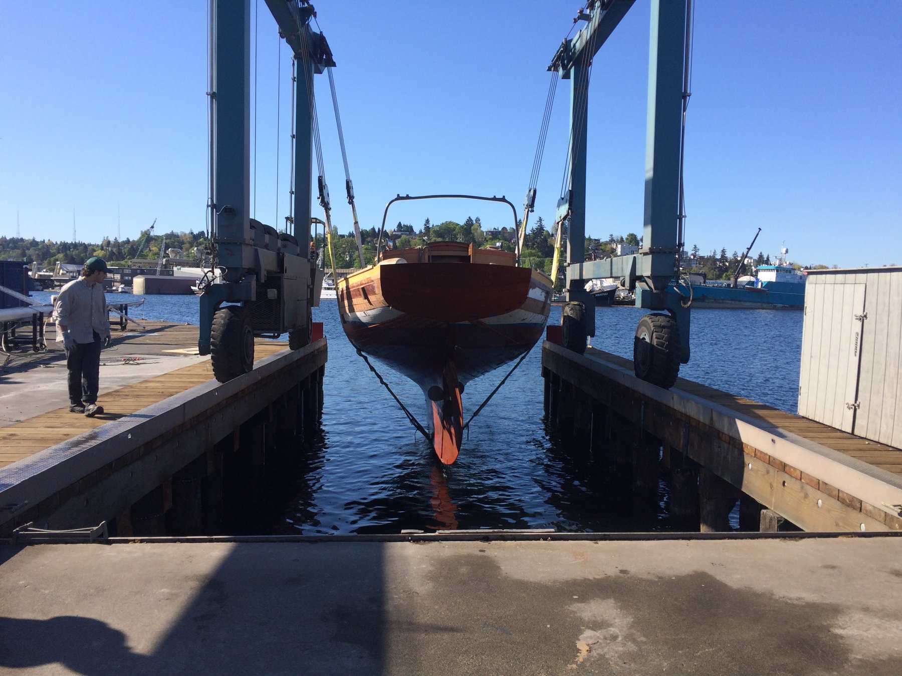 The classic 1946 Philip Rhodes-designed Merry Maiden suspended in the boat lift at a Pacific Northwest shipyard, her gleaming wooden hull poised between sea and sky in this serene moment of maintenance.