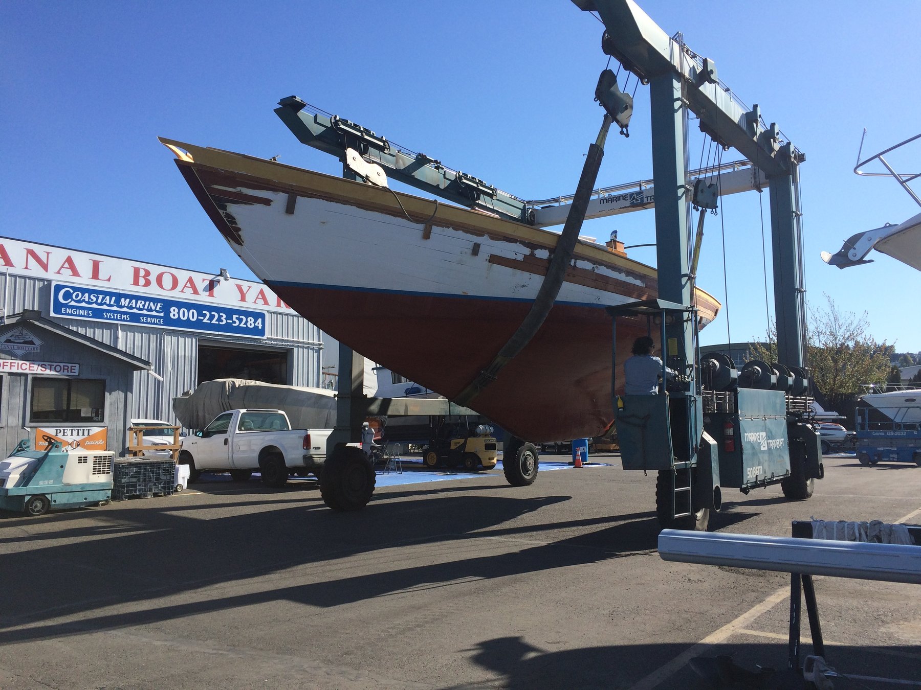 The classic 1946 Philip Rhodes-designed Merry Maiden suspended in the travel lift at a Puget Sound boatyard, her elegant hull exposed under the clear Pacific Northwest sky.