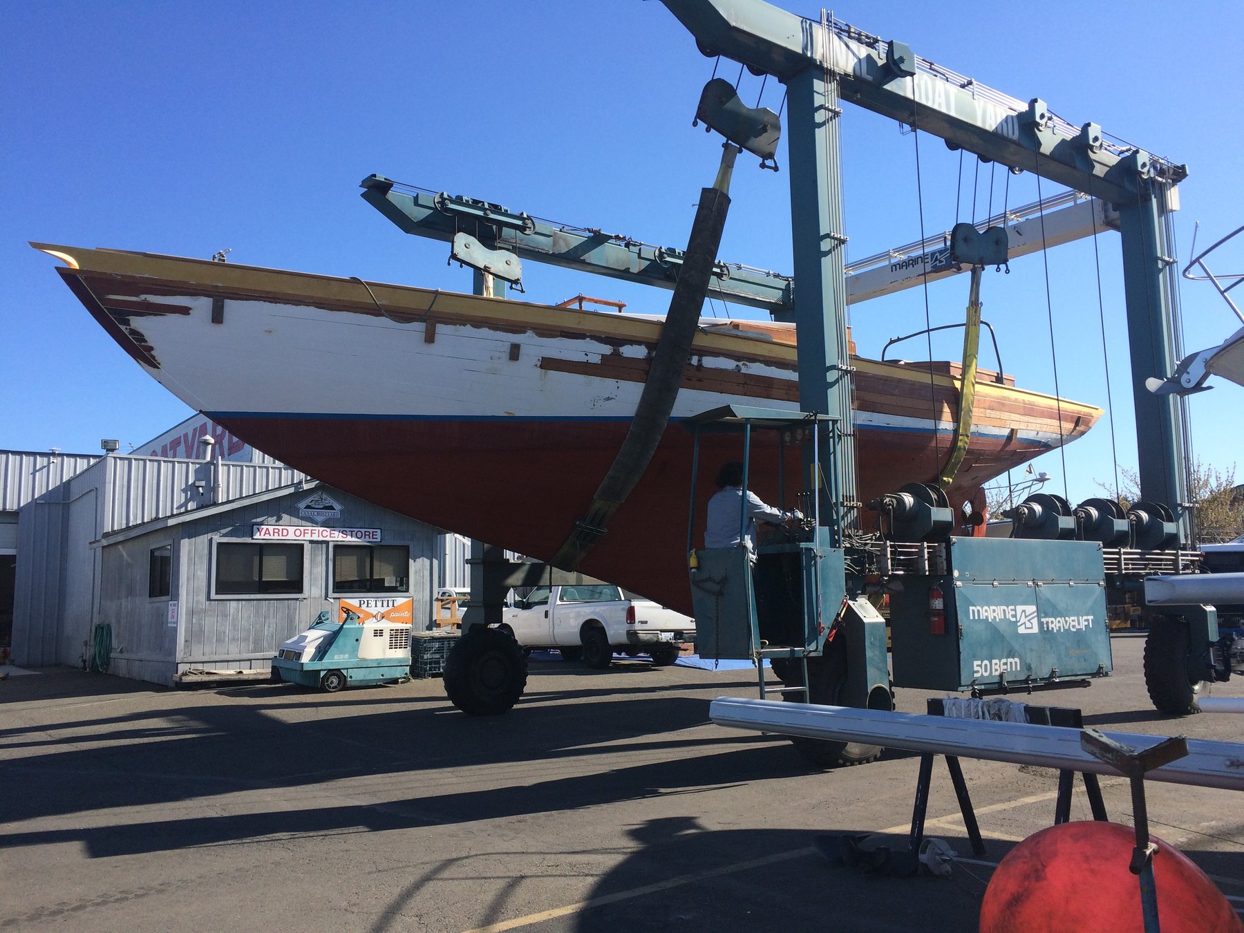 The classic 1946 Philip Rhodes-designed Merry Maiden rests in a Pacific Northwest boatyard, suspended by a travel lift against a clear spring sky, awaiting maintenance or restoration.