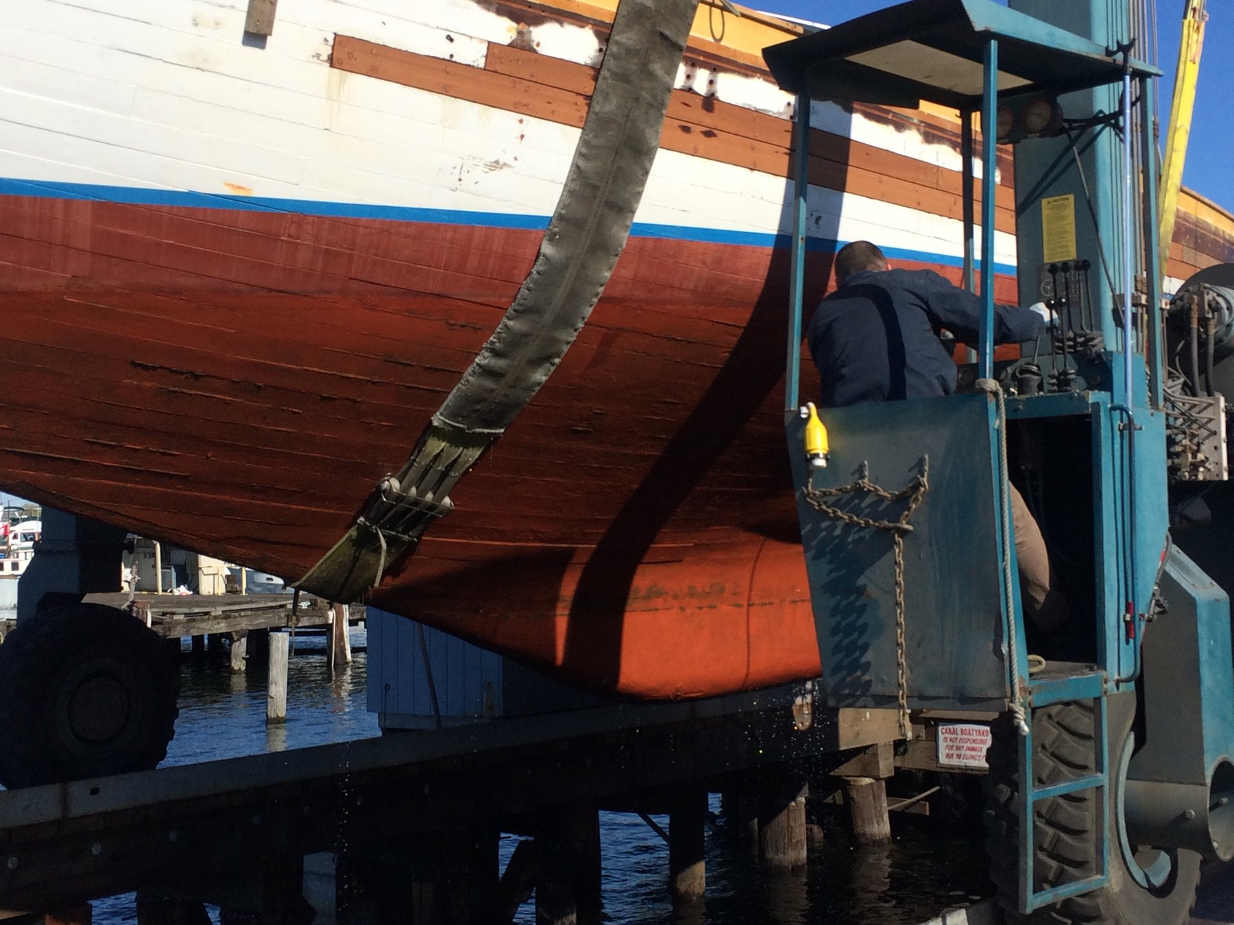 A worker tends to the classic 1946 Philip Rhodes-designed Merry Maiden during haul-out at a Puget Sound boatyard, her distinctive red hull and white topsides gleaming in the afternoon sun.