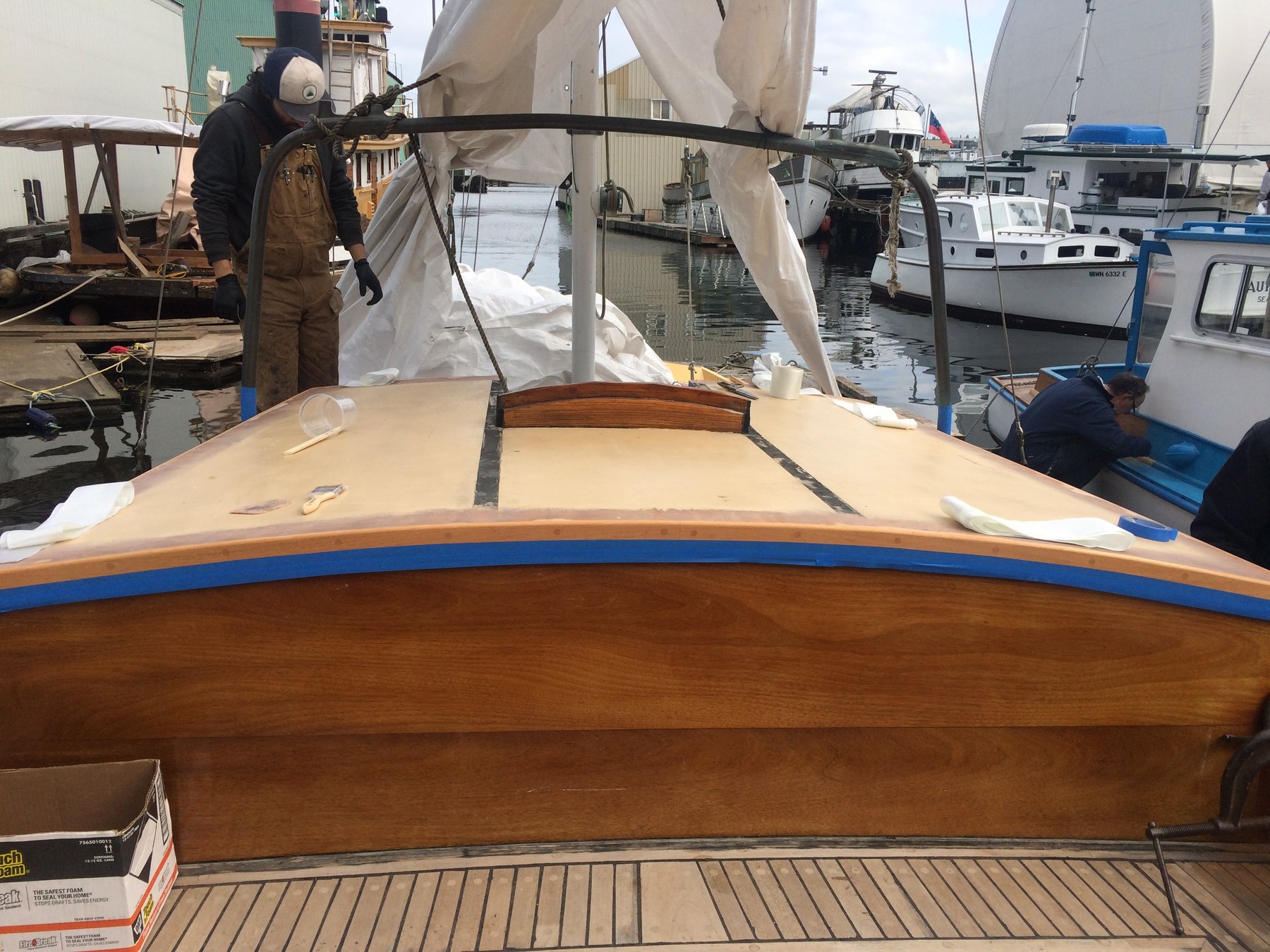 A craftsman carefully works on the bright teak deck of the classic 1946 Philip Rhodes design Merry Maiden, her canvas canopy protecting the meticulous restoration work underway in a Puget Sound boatyard.