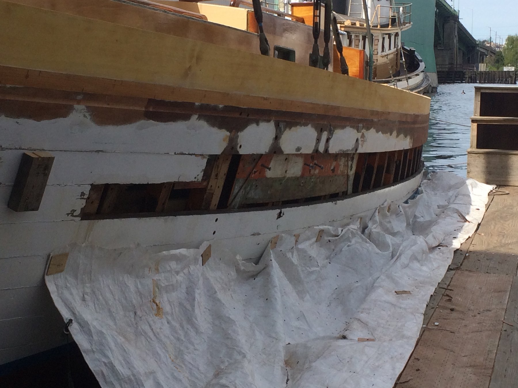 The Merry Maiden's hull reveals extensive structural work during restoration, with protective coverings draping her classic wooden frame in the shipyard.