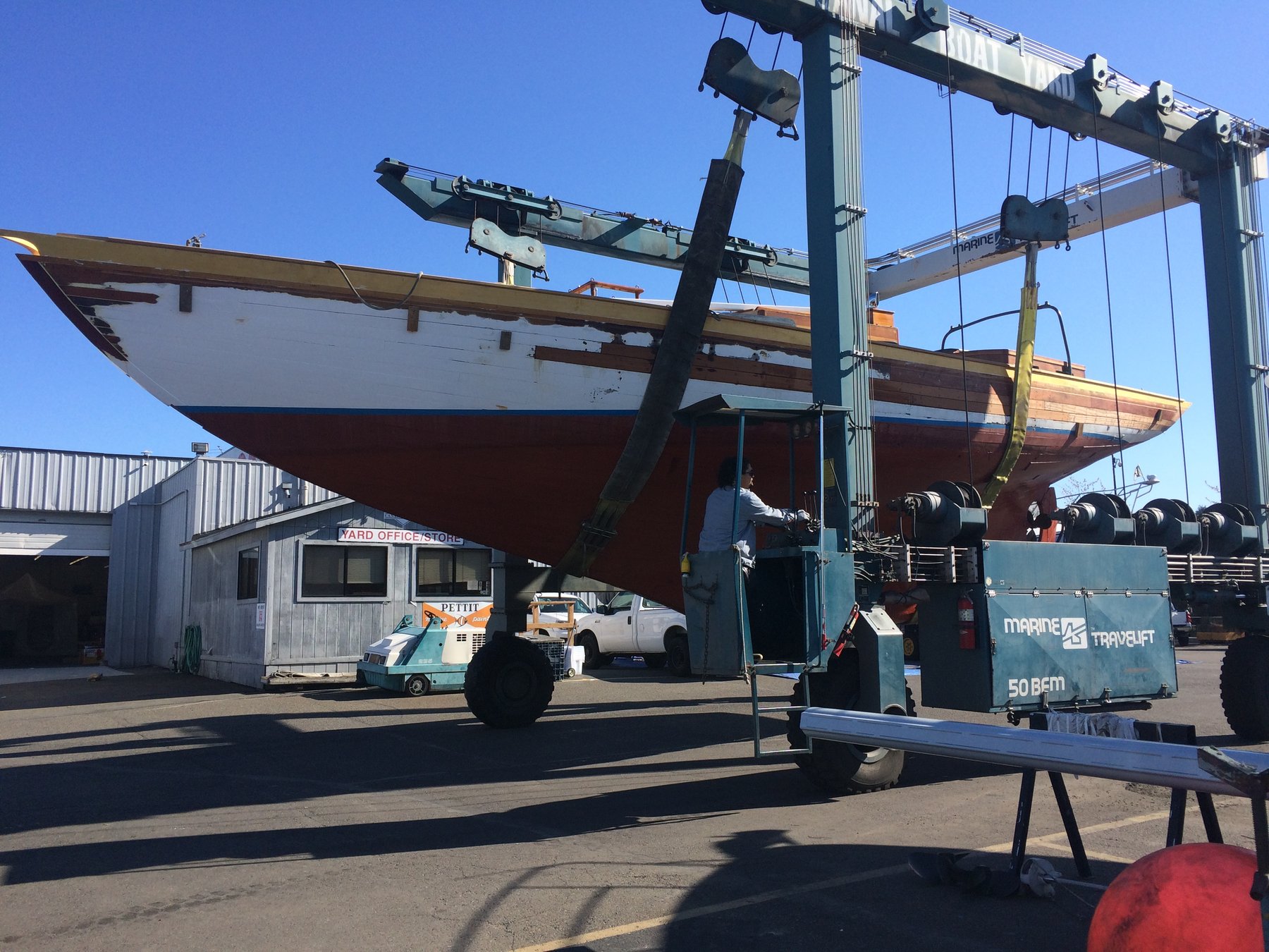 The classic 1946 Philip Rhodes-designed Merry Maiden suspended in the travel lift at a Pacific Northwest boatyard, her elegant wooden hull exposed and ready for maintenance.